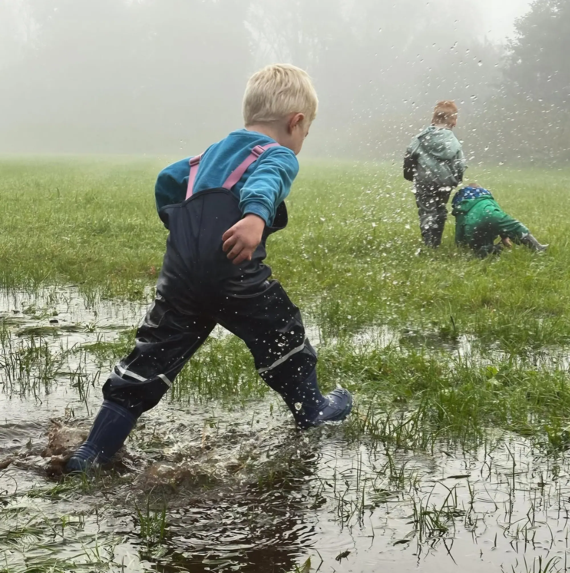 Kinderen spelen buiten in regenlaarzen in een plas op een mistig grasveld